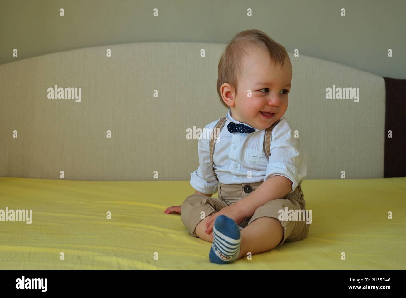 Niedliches kleines Baby, das mit Hemd und Fliege auf dem Bett sitzt Stockfoto