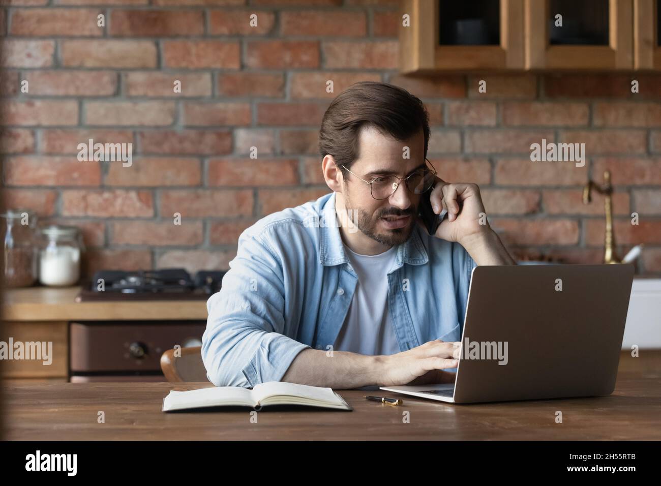 Fokussierter Millennial-Typ arbeitet an einem Laptop, der in Telefongespräche verwickelt ist Stockfoto