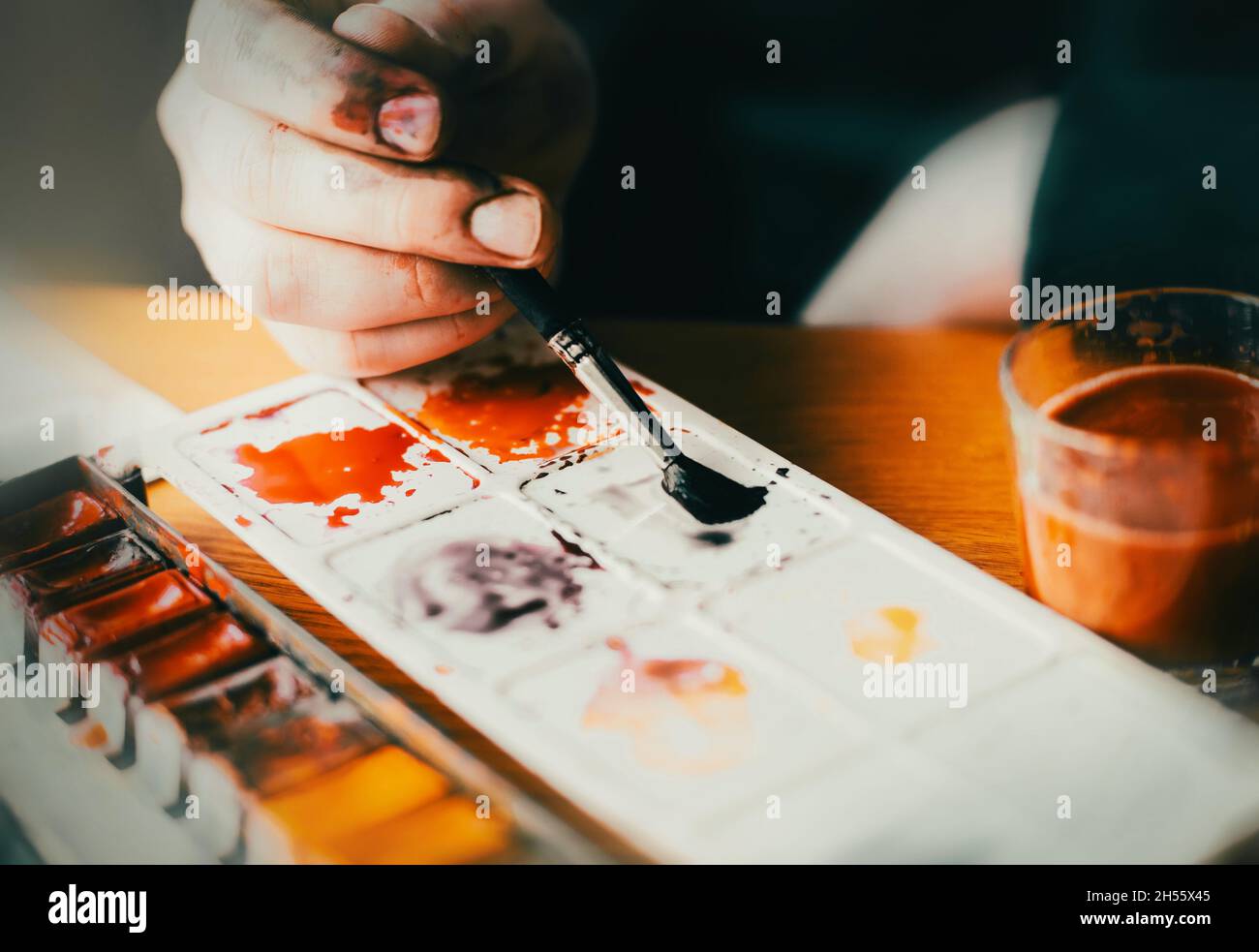 Ein Künstler in einer dunklen Jacke hält einen Pinsel in der Hand, mit dem er helle Aquarellfarben auf einer Palette mischt. Der Prozess des Zeichnens. Kreativi Stockfoto