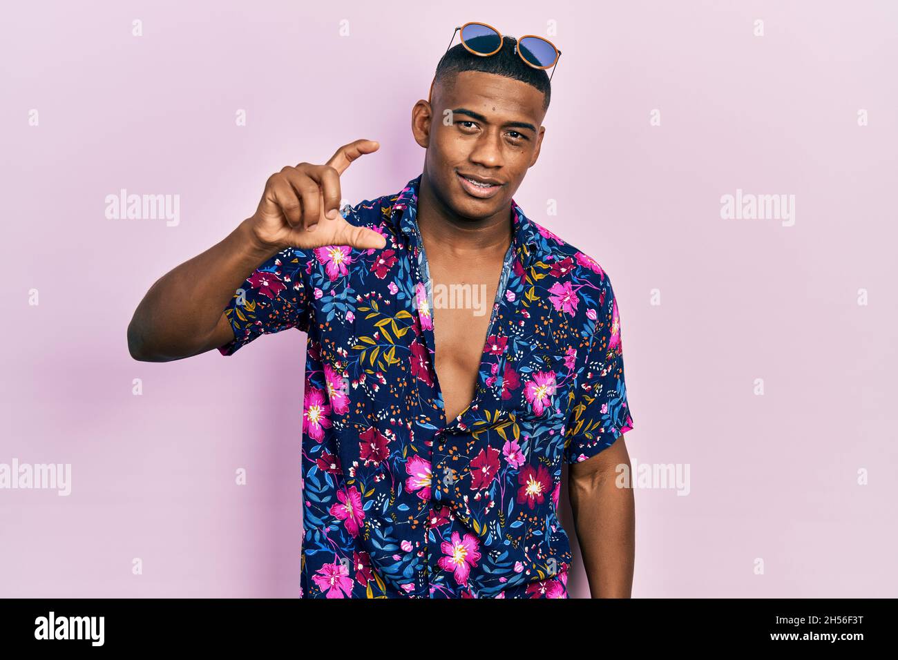 Junger schwarzer Mann mit hawaiiihemd und Sonnenbrille, lächelnd und selbstbewusst gestikulend, mit der Hand und einem kleinen Schild mit den Fingern und der Kamera Stockfoto