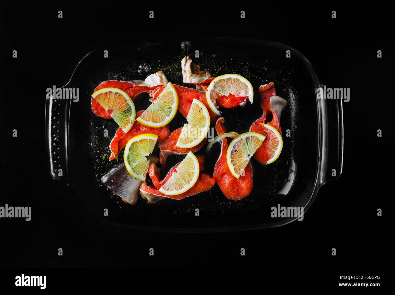 Rote Fischstücke mit Zitrone und Gewürzen in einer Glasschale auf schwarzem Hintergrund. Stockfoto