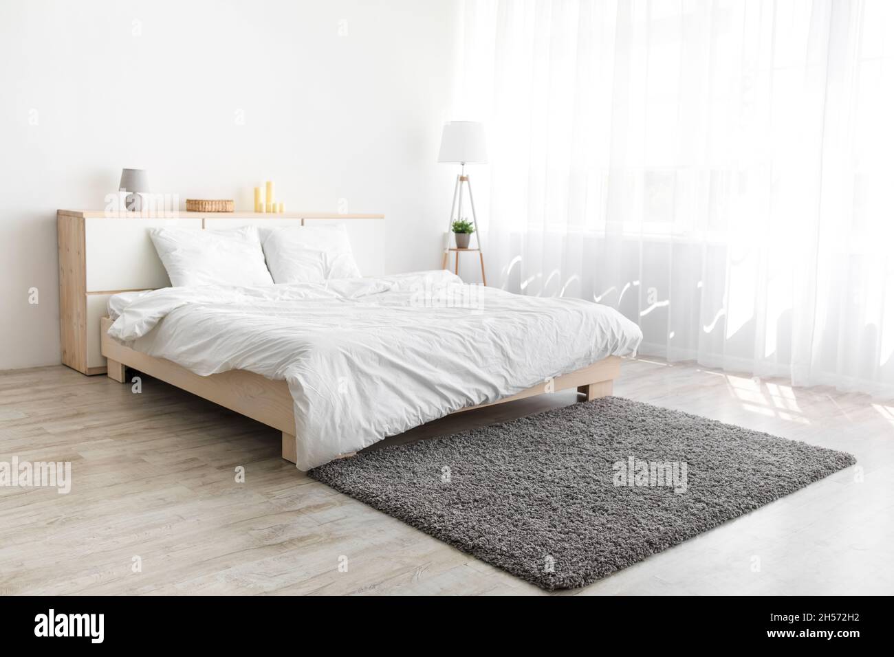 Großes Bett mit bequemer Bettwäsche, weicher grauer Teppich, Lampe auf dem Boden, Möbel mit Accessoires Stockfoto