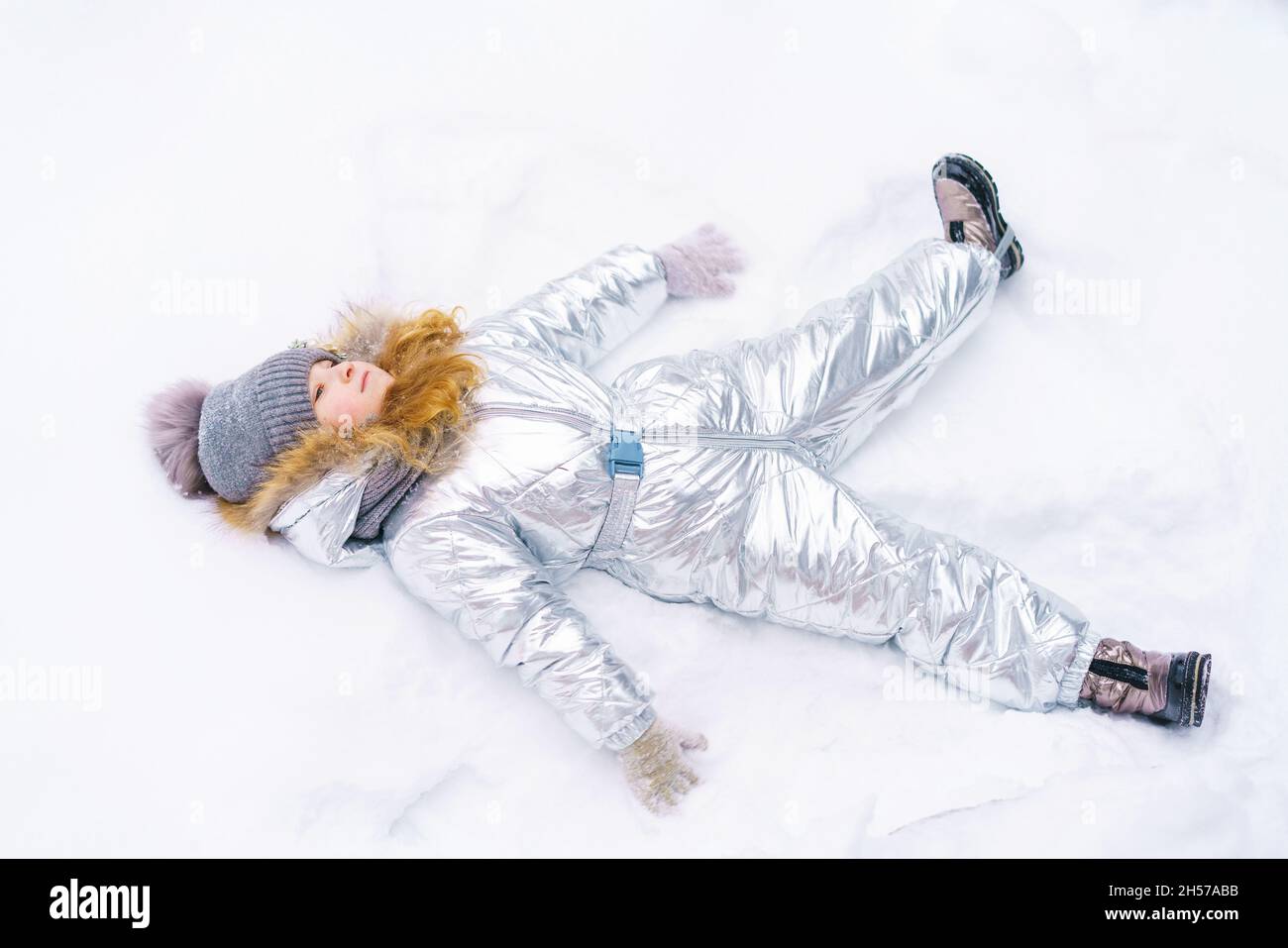 Mädchen spielt im Schnee. Das Kind liegt im Schnee und breitet seine Arme in Form eines Engels zur Seite aus. Glückliches kleines Mädchen in warmem silbernem Jumpsuit Stockfoto
