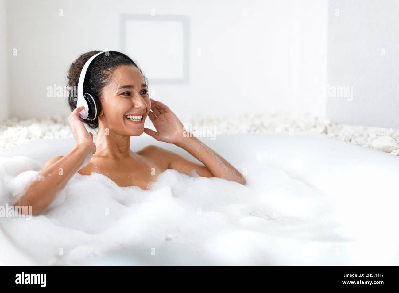 Glückliche junge Dame mit Kopfhörern, die sich im schäumenden Bad entspannt, zu Hause Musik hört, freien Platz Stockfoto