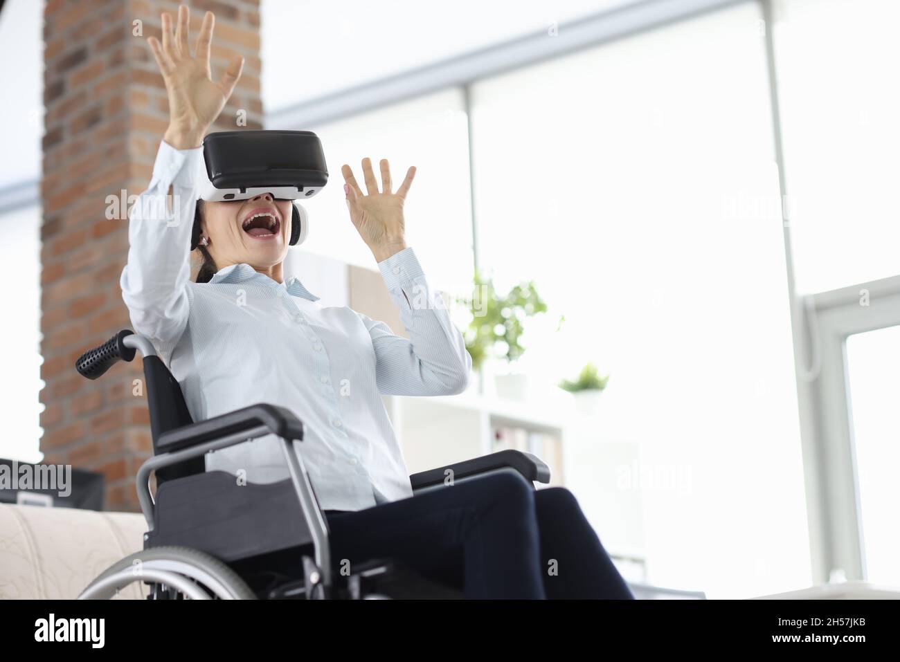 Frau, die im Rollstuhl sitzt und eine Virtual-Reality-Brille trägt und ihre Hände winkt Stockfoto