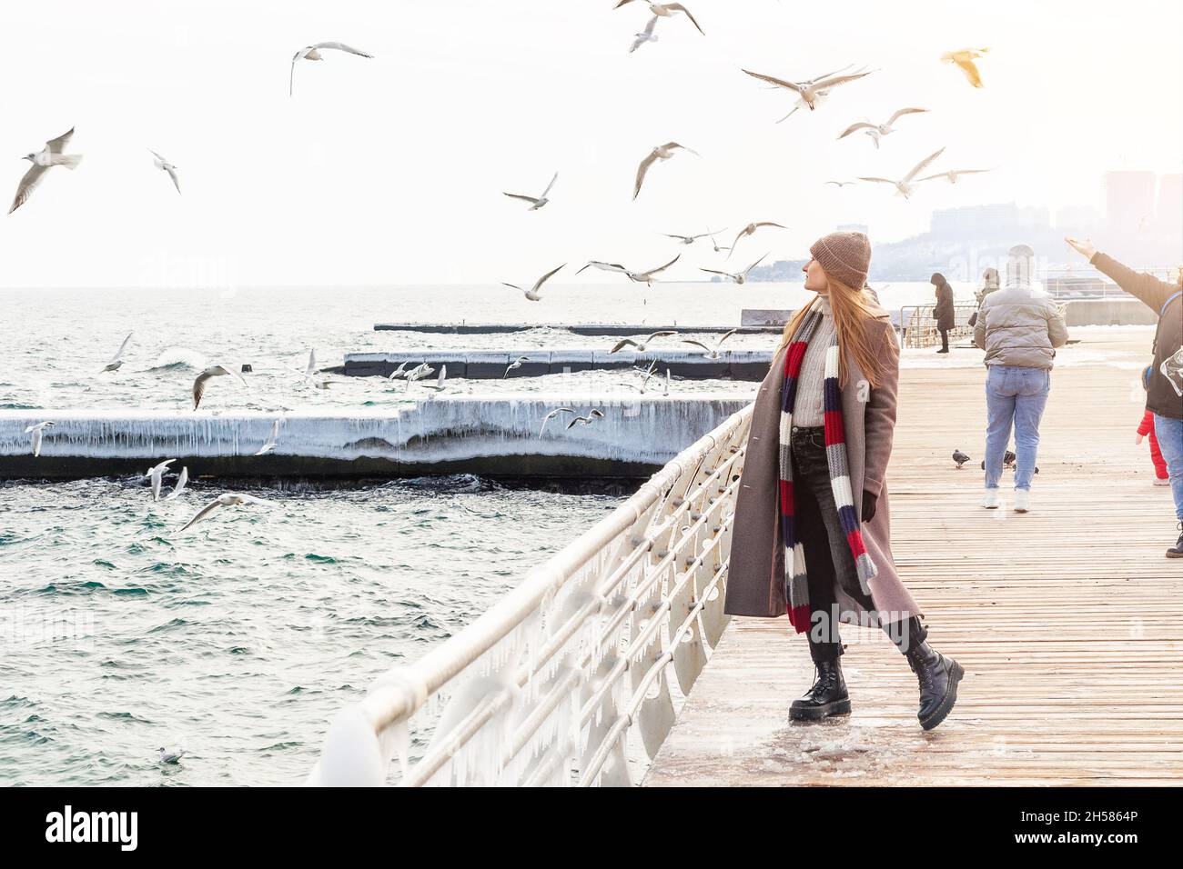 Wanderfrau auf dem Pier im Winter. Stilvolle Dame trägt Wintermantel, raue Stiefel, Schal und Hut allein in der Nähe des Meeres stehen Stockfoto
