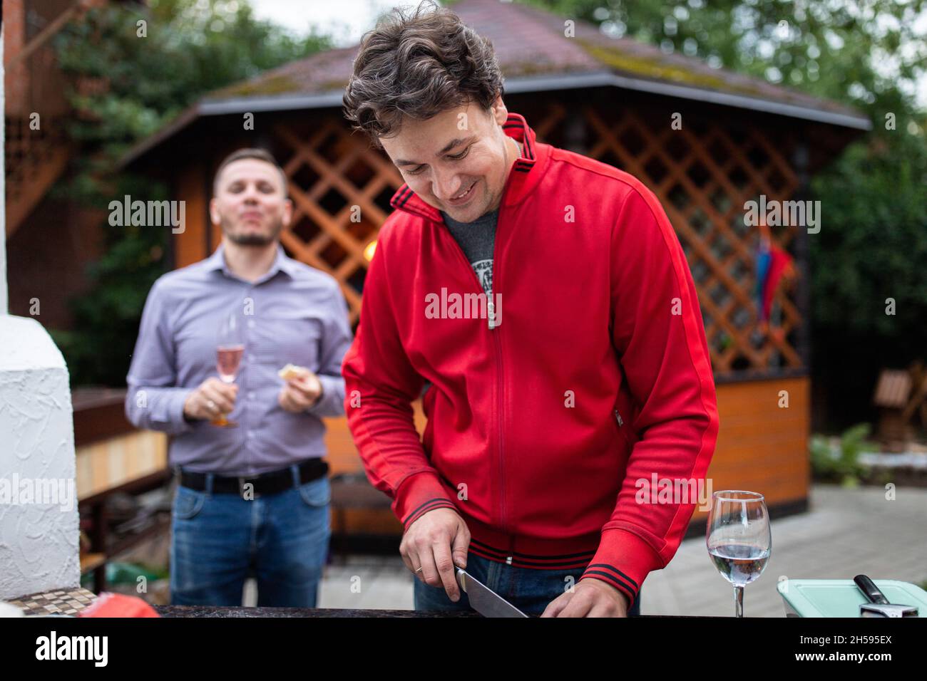 Ein Mann bereitet auf einer Hinterhofparty Essen für seine Freunde zu. Er schneidet Pizzaprodukte, im Hintergrund trinkt ein Freund Champagner. Stockfoto