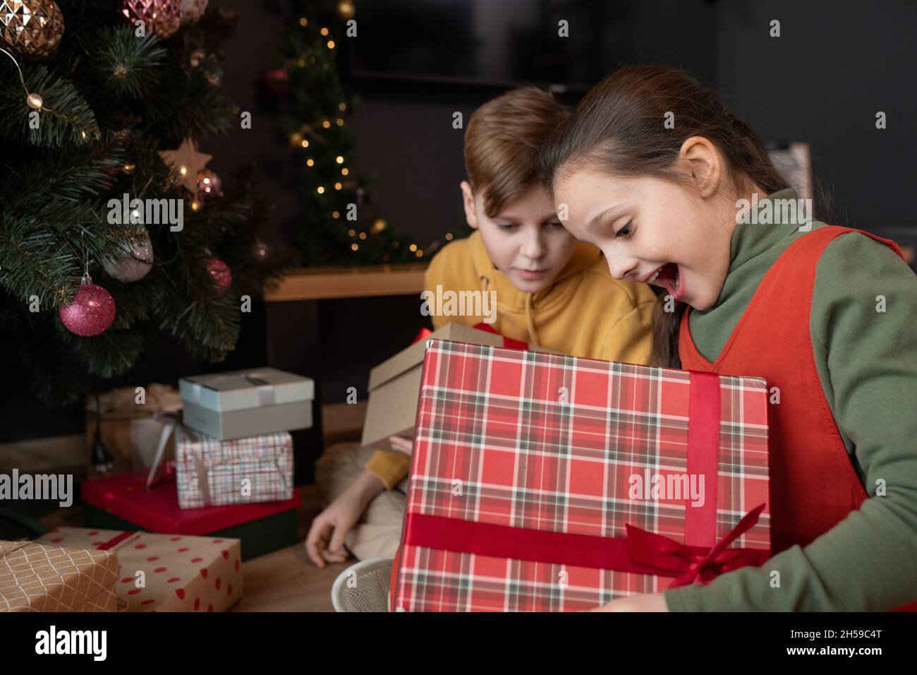 Aufgeregt hübsches Mädchen mit offenem Mund sitzt auf dem Boden mit dekorierten Baum und Eröffnung Weihnachtsgeschenk mit Bruder Stockfoto