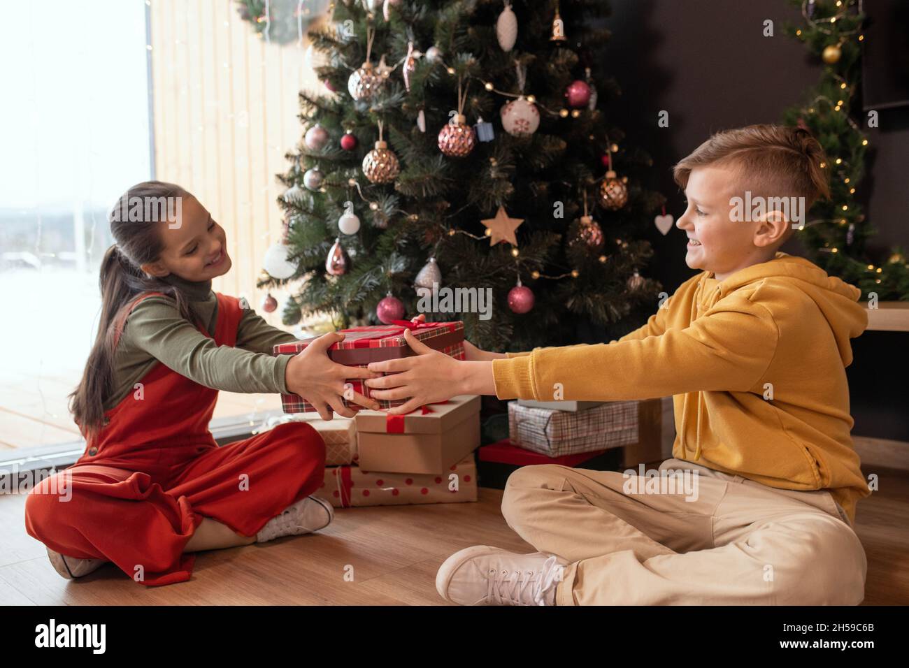 Lächelnder Teenager-Junge in gelbem Pullover, der mit gekreuzten Beinen auf dem Boden in der Nähe des Weihnachtsbaums sitzt und der Schwester Weihnachten vorgibt Stockfoto