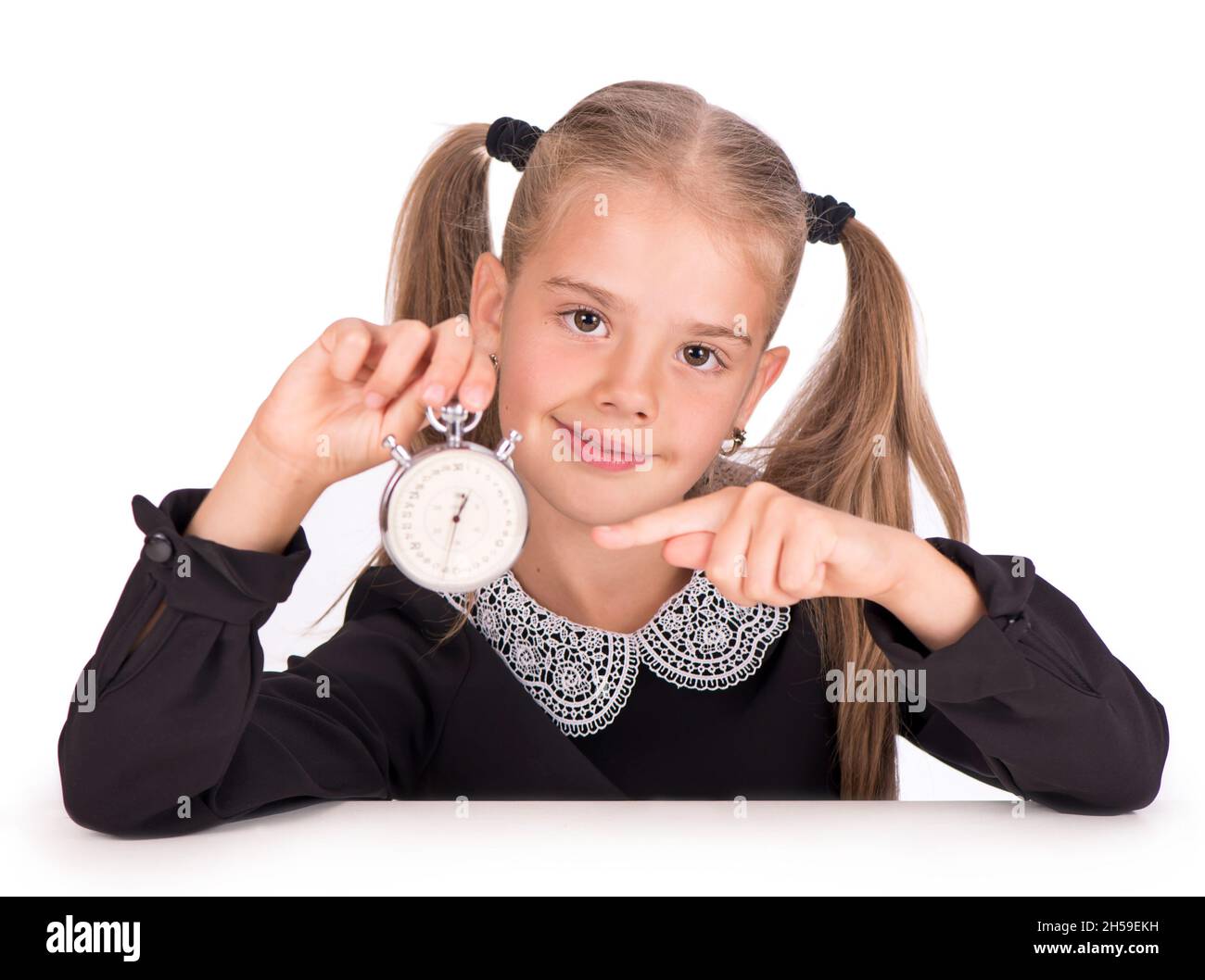 Das kleine Mädchen, blond in einem Schulkleid, weist auf eine Stoppuhr auf weißem Hintergrund hin Stockfoto