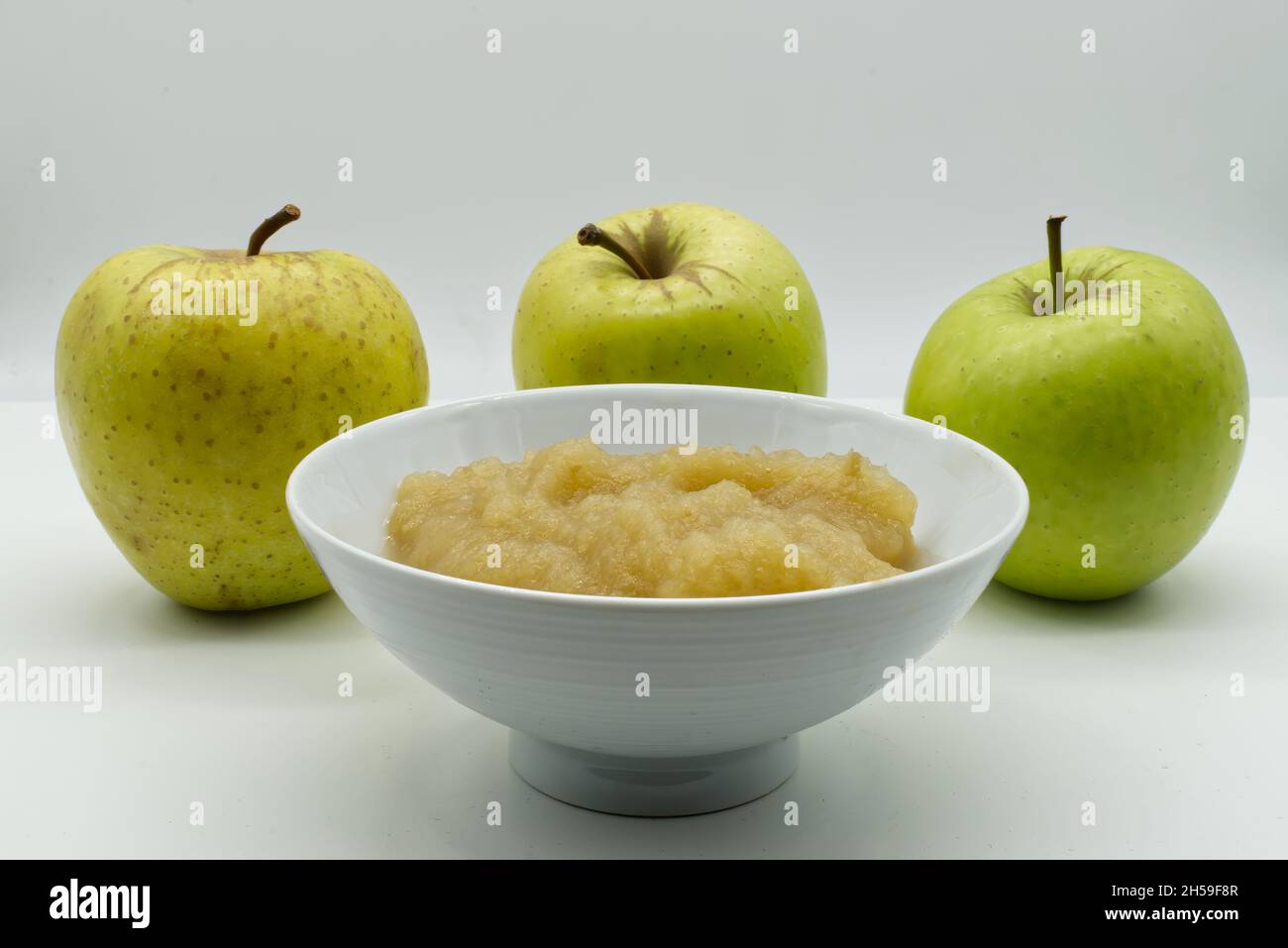 Apfelsauce in einer weißen Schüssel mit rohen Äpfeln auf dem Hintergrund. Apfelmus und frische grüne Äpfel isoliert auf weißem Hintergrund. Stockfoto