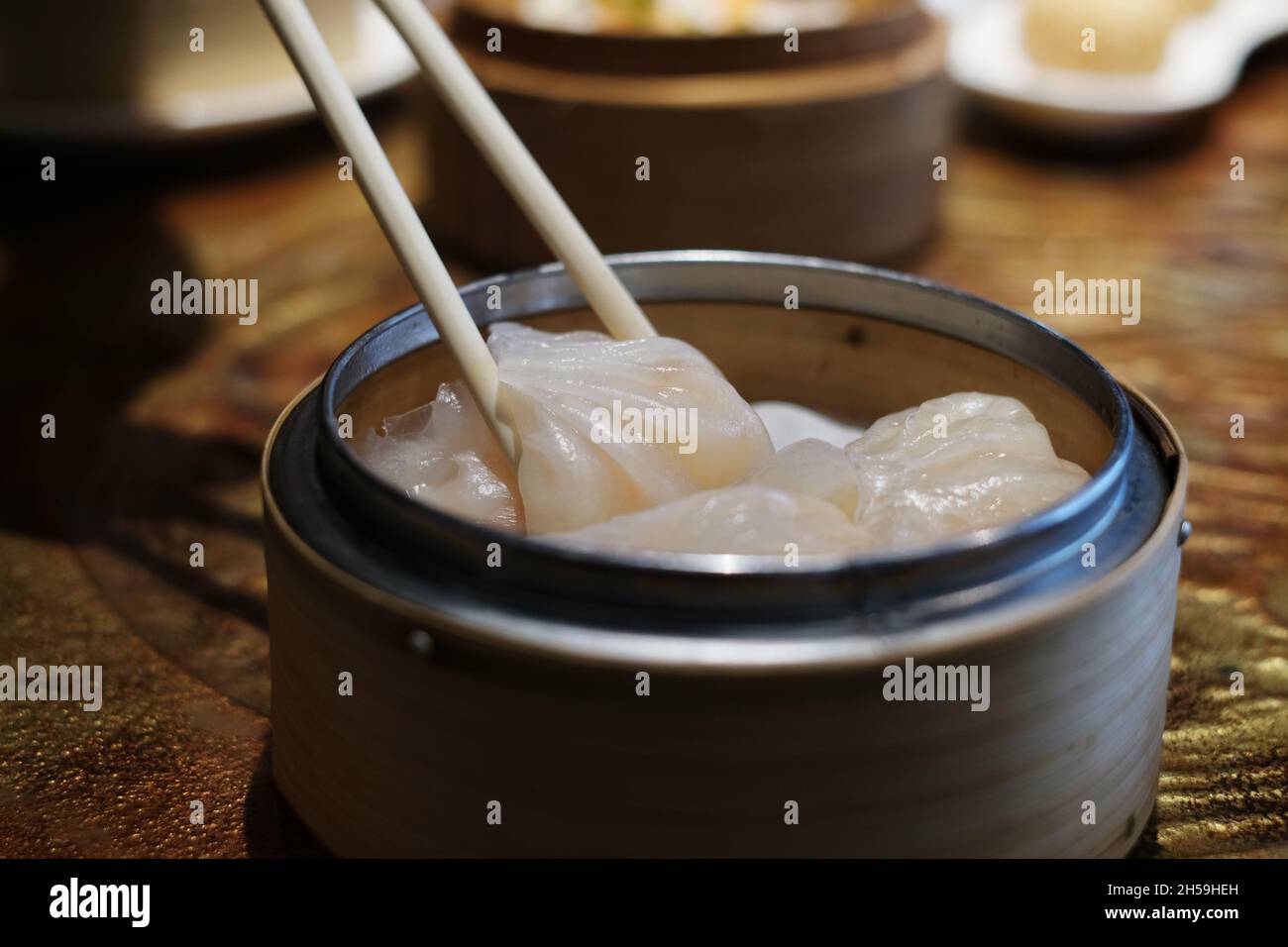 Ein Nahaufnahme-Bild von Bambus-Essstäbchen, die weiße chinesische gefüllte gedämpfte Knödel aufsammeln, auch bekannt als Dim Sum aus einem runden Bambuskorb auf einem Stockfoto