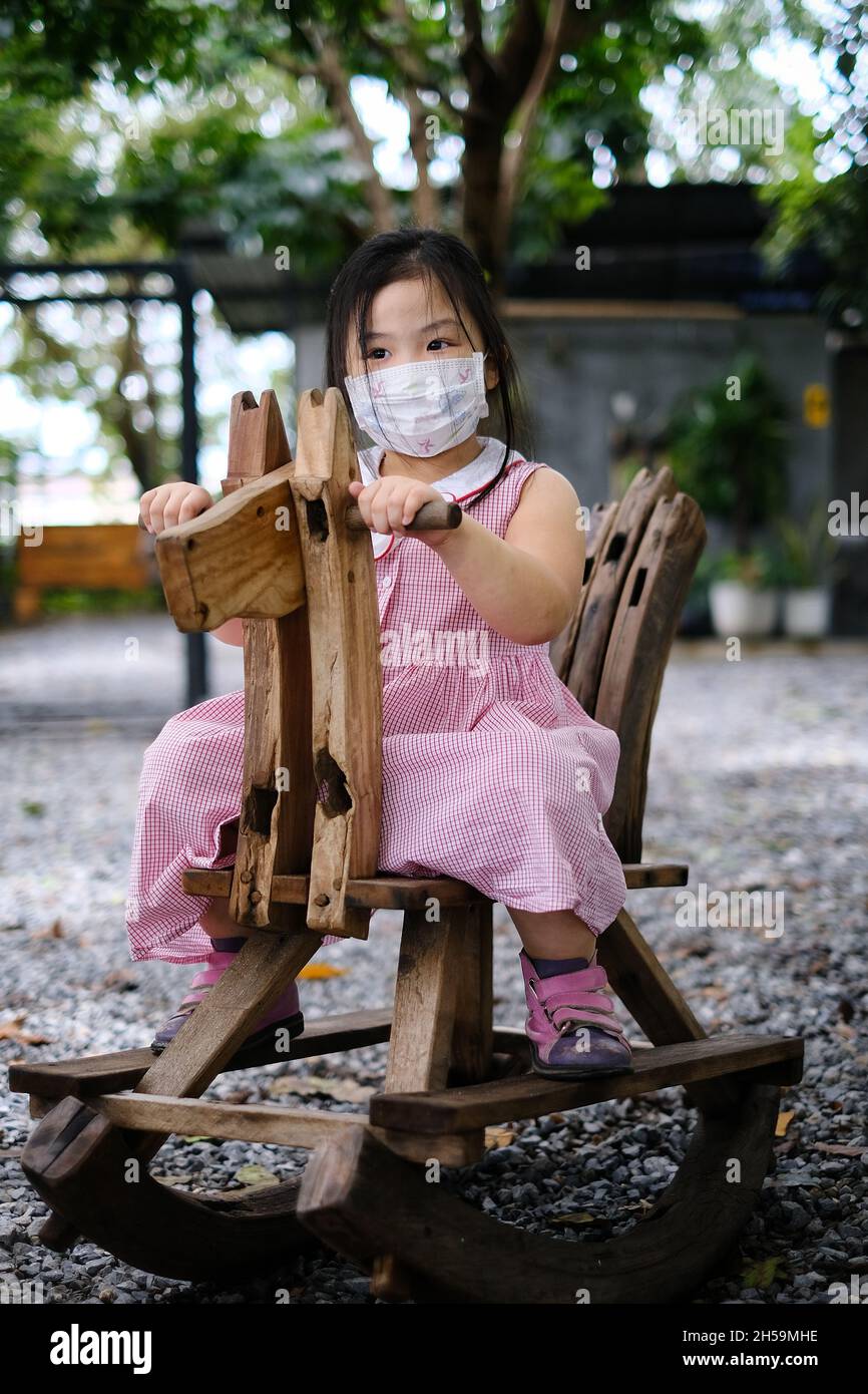 Ein süßes junges asiatisches Mädchen mit einer Gesichtsmaske über ihrem Mund reitet auf einem pferdeförmigen hölzernen Schaukelstuhl auf einem Spielplatz während Covid-19 pand Stockfoto