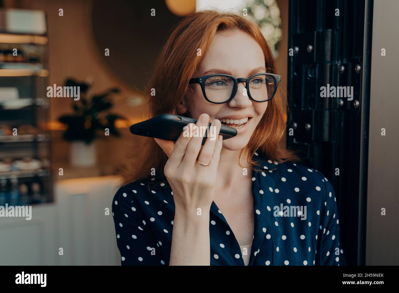 Eine fröhlich lächelnde Geschäftsfrau spricht am Lautsprecher über das Handy, während sie den Coffee Shop verlässt Stockfoto