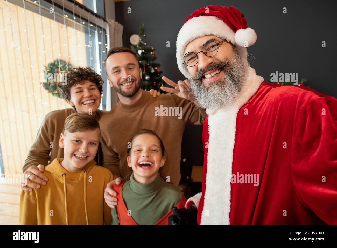 Selfie vom glücklichen Weihnachtsmann in Mütze und Brille, posiert mit positiver Familie gegen den Weihnachtsbaum zu Hause Stockfoto