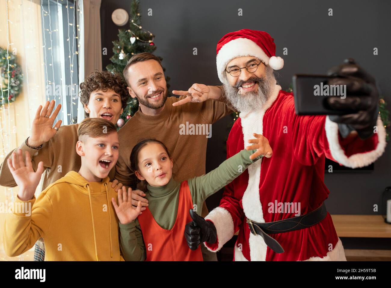 Positiver Weihnachtsmann in Brillen, der während der Weihnachtszeit mit fröhlicher Familie telefoniert und Daumen nach oben zeigt Stockfoto