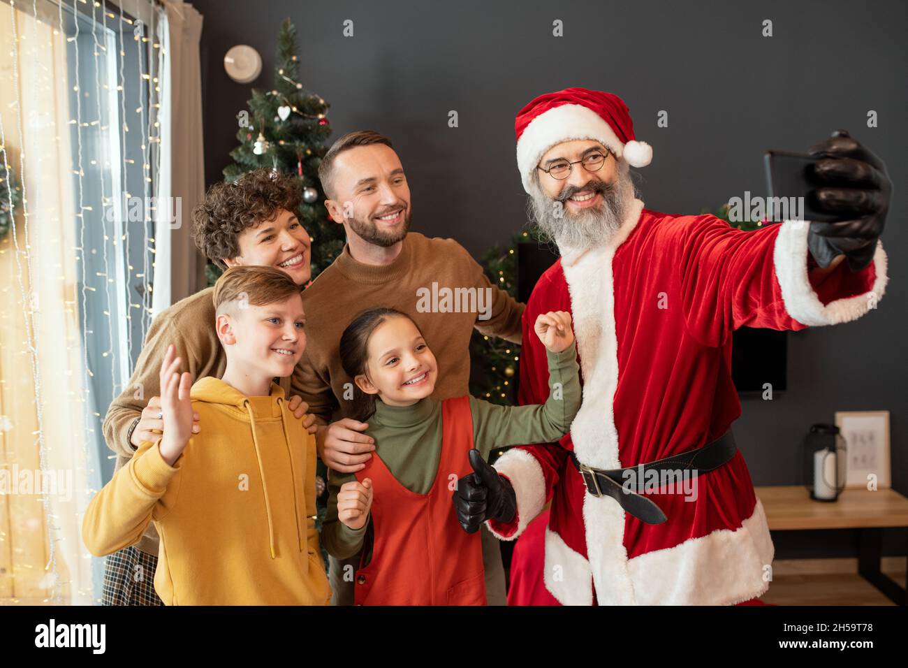 Lächelnder Weihnachtsmann im roten Kostüm, Selfie mit glücklicher Familie auf dem Smartphone gegen den Weihnachtsbaum Stockfoto