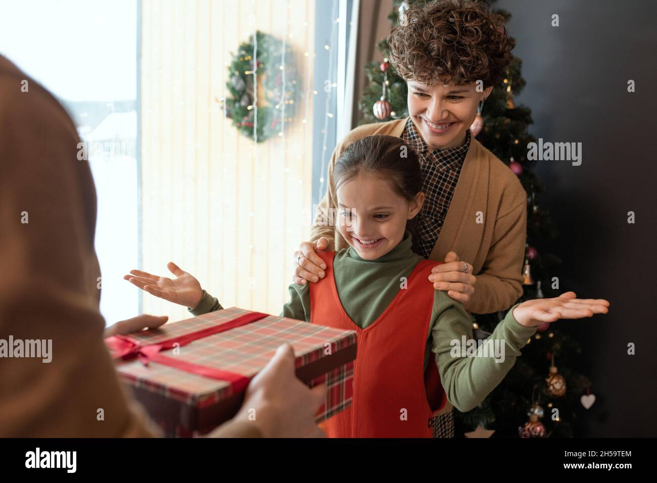 Lächelnde Mutter hält die Hände auf der Schulter der Tochter, während der Vater ihr im Wohnzimmer ein Weihnachtsgeschenk gibt Stockfoto
