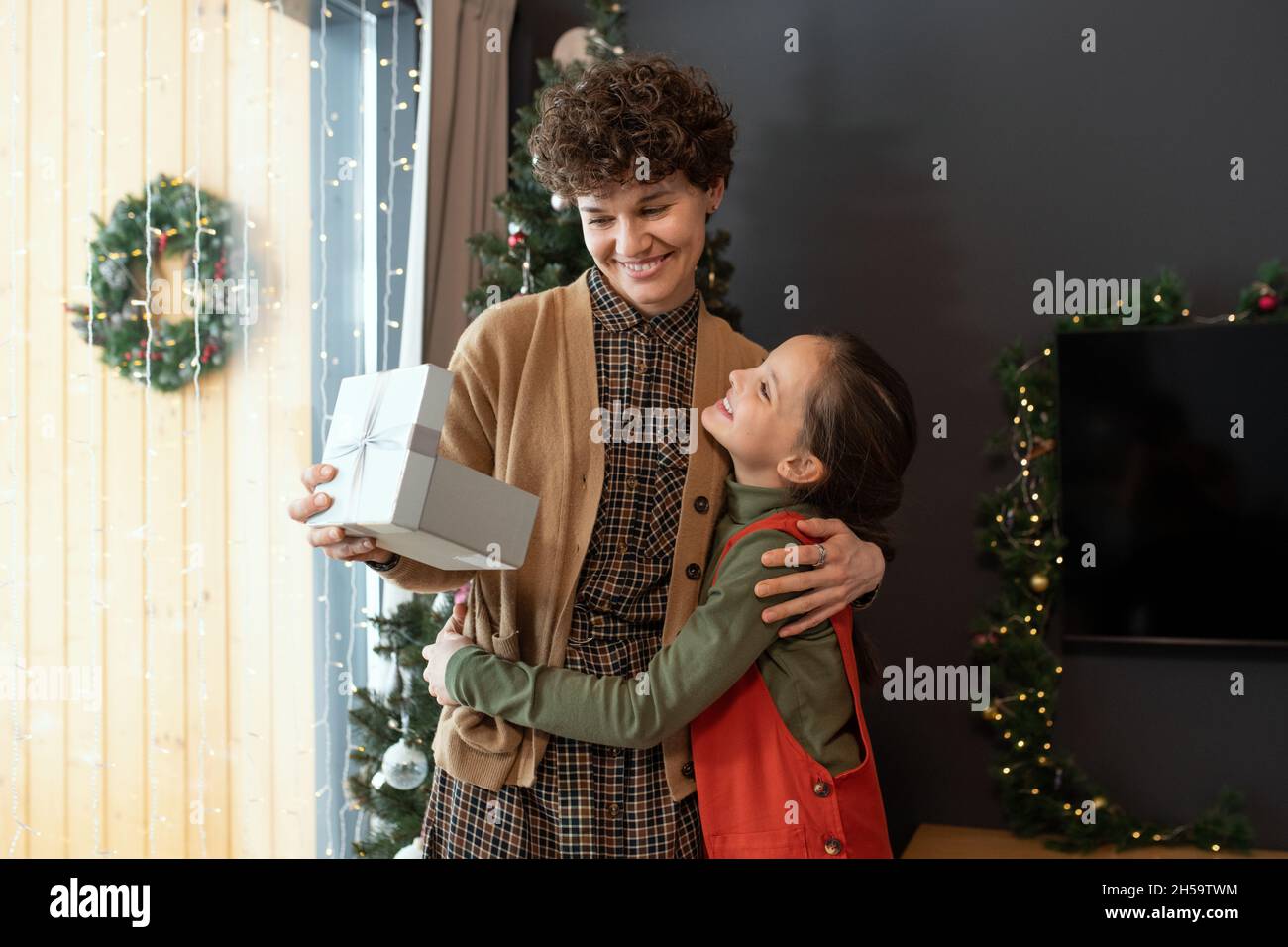 Lächelnde, lockige Frau umarmt die Tochter, während sie zu Hause Weihnachtsgeschenk von ihr erhält Stockfoto