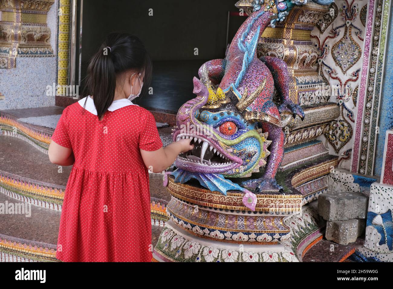 Bangkok / Thailand - 2021/11/01: Die Rückansicht eines jungen asiatischen Mädchens in rotem Kleid berührt die Zähne eines farbenfrohen thailändischen Fabelwesens namens Mo Stockfoto