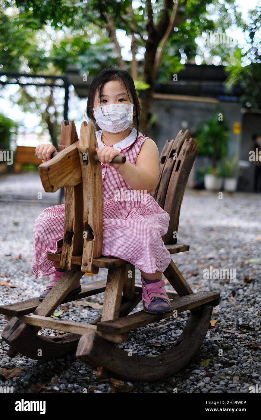 Ein süßes junges asiatisches Mädchen mit einer Gesichtsmaske über ihrem Mund reitet auf einem pferdeförmigen hölzernen Schaukelstuhl auf einem Spielplatz während Covid-19 pand Stockfoto