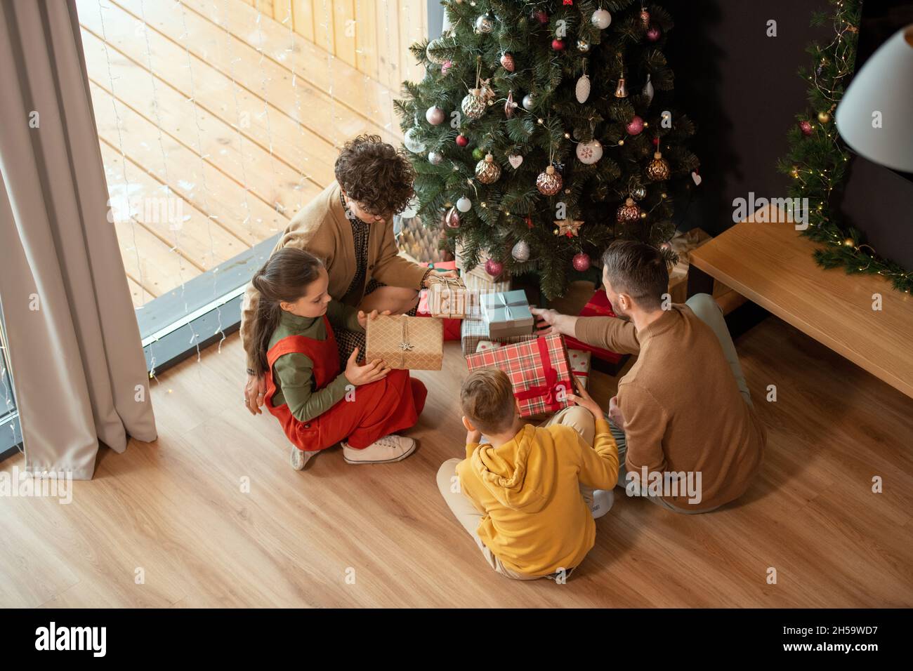 Direkt über der Ansicht der Familie mit zwei Kindern, die um den geschmückten Baum sitzen und am Weihnachtsmorgen Geschenke austauschen Stockfoto