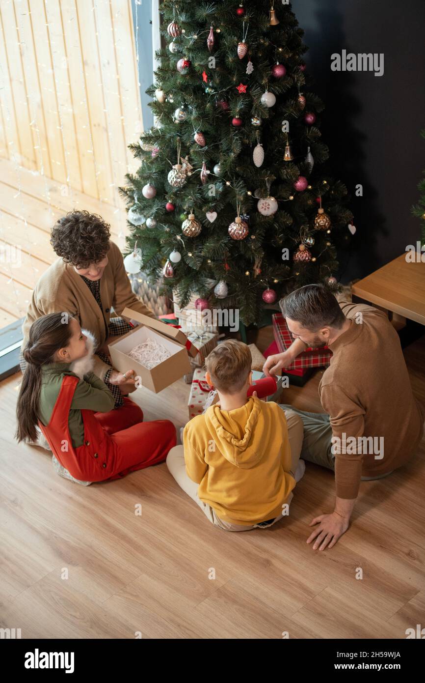 Eltern sitzen an geschmückten Bäumen und Geschenkschachteln darunter und schenken Kindern Weihnachtsgeschenke Stockfoto
