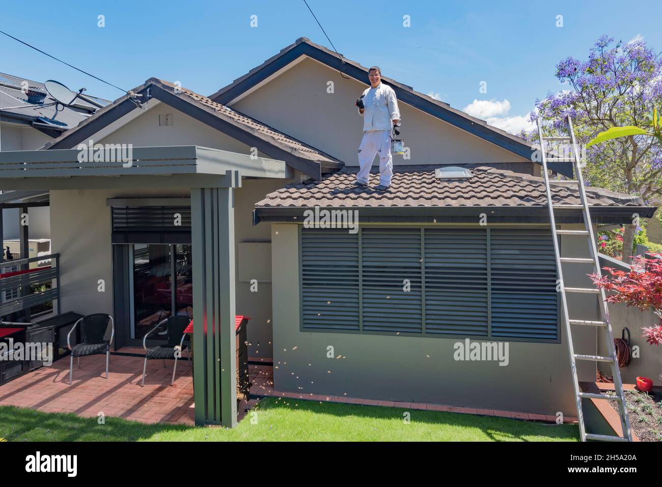 Ein Mann in weißen Overalls steht mit einem Pinsel in der Hand auf seinem Hausdach, während seine Honigbienen aus seinem Flow Hive um seinen Hinterhof in Sydney fliegen Stockfoto