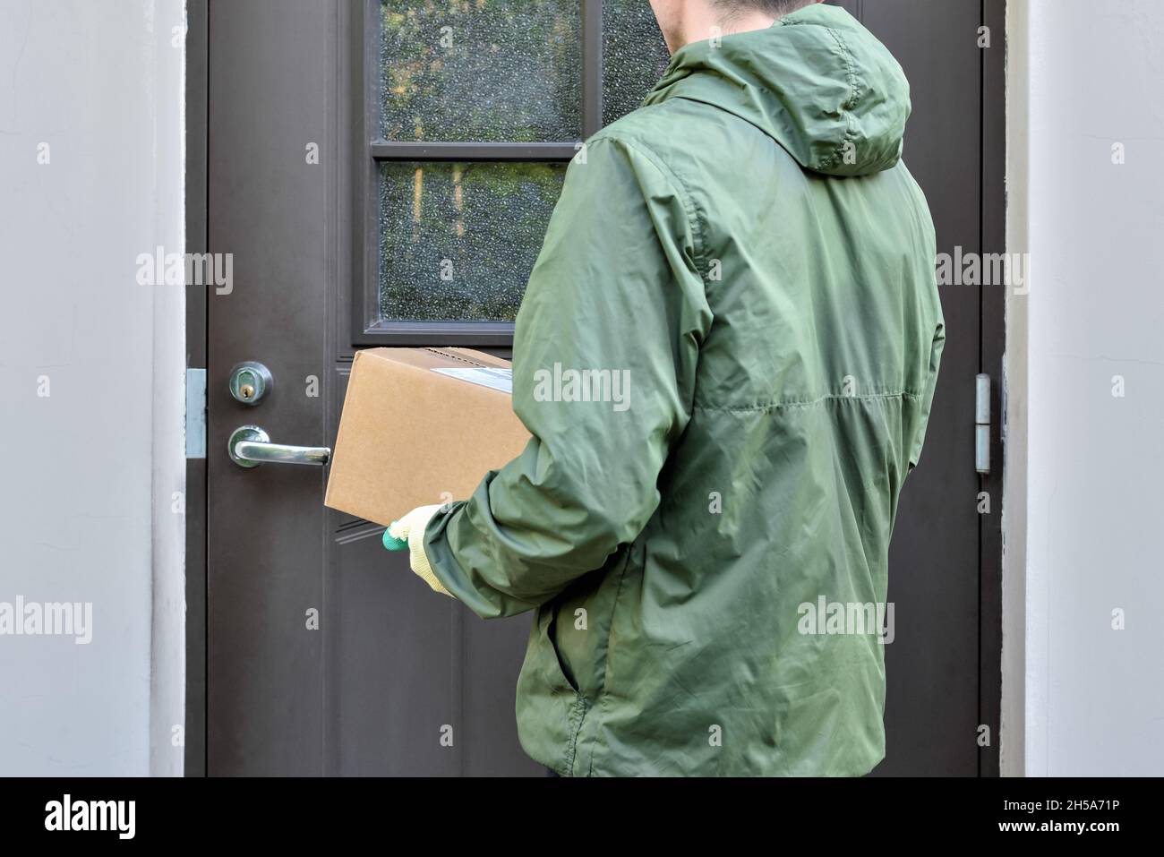 Paketkarton in den Händen eines Postboten, der vor der Tür des Hauses steht Stockfoto