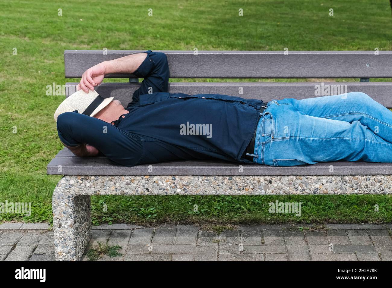 Ein Mann ruht auf einer Bank in einem öffentlichen Stadtpark und bedeckt seinen Kopf mit einem Hut. Das Konzept des Neustarts, der Veränderung oder des Rücksetzens Ihres Lebens und Makins Stockfoto