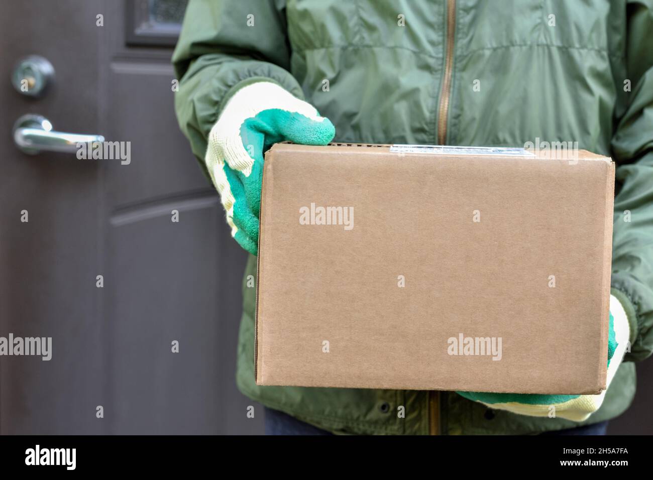 Paketkarton in den Händen des Postboten auf dem Hintergrund der Tür des Hauses. Lieferung des Paketkurierdienstes zu Ihnen nach Hause. Stockfoto