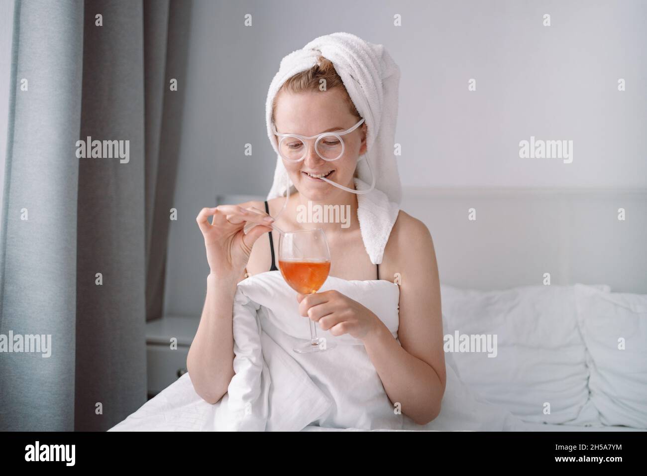 Glückliche Frau mit Handtuch auf dem Kopf liegt im Bett unter Decke isoliert über weißen Wand Hintergrund trinken Wein, haben einen Ventilator. Alkohol, Feiern und Trauern Stockfoto