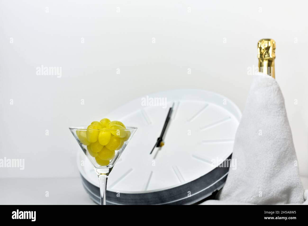 Weinglas mit Trauben auf dem Hintergrund einer verschwommenen Uhr und einer Flasche Champagner. Spanische Tradition, zwölf Trauben zu essen, um Neujahr zu feiern. Stockfoto