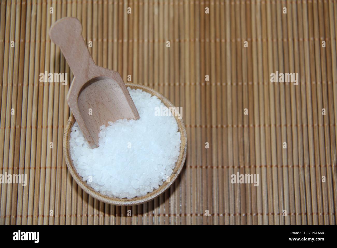 Grobes Salz, in einer Holzschüssel, mit einem Messlöffel, auf einer Bambusmatte. Blick von oben. Stockfoto