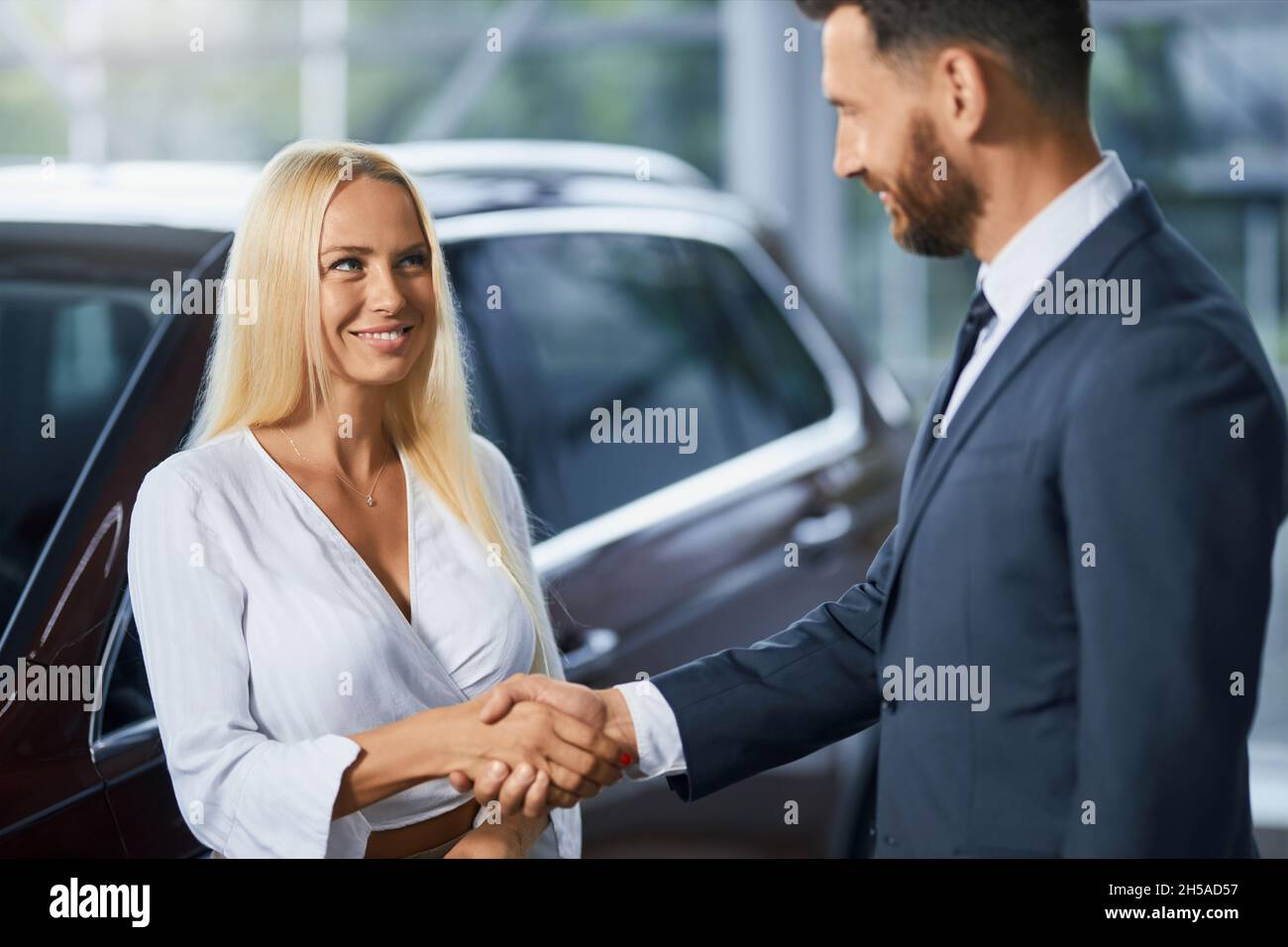 Kaukasischer Autohändler in stilvollem Anzug Hände schütteln mit charmanten Frau mit blonden Haaren nach erfolgreicher Vereinbarung des Kaufs Auto. Konzept von Menschen, Verkauf und Kauf. Stockfoto