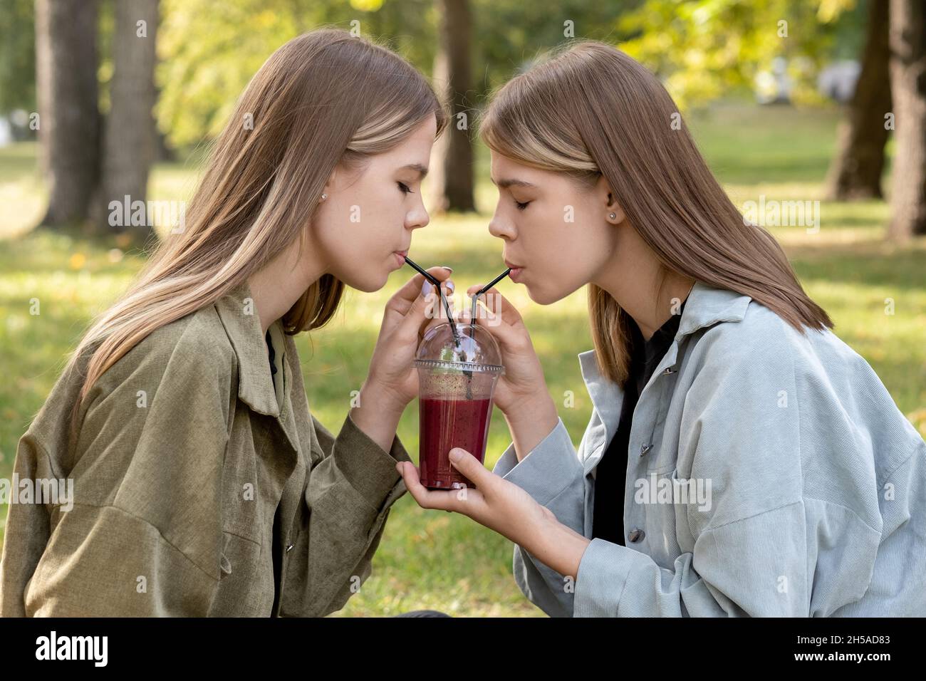 Seitenansicht von Zwillingsmädchen in Casualwear, die ein großes Glas frischen Obst-Smoothie teilen, während sie den Sommertag im Park verbringen Stockfoto