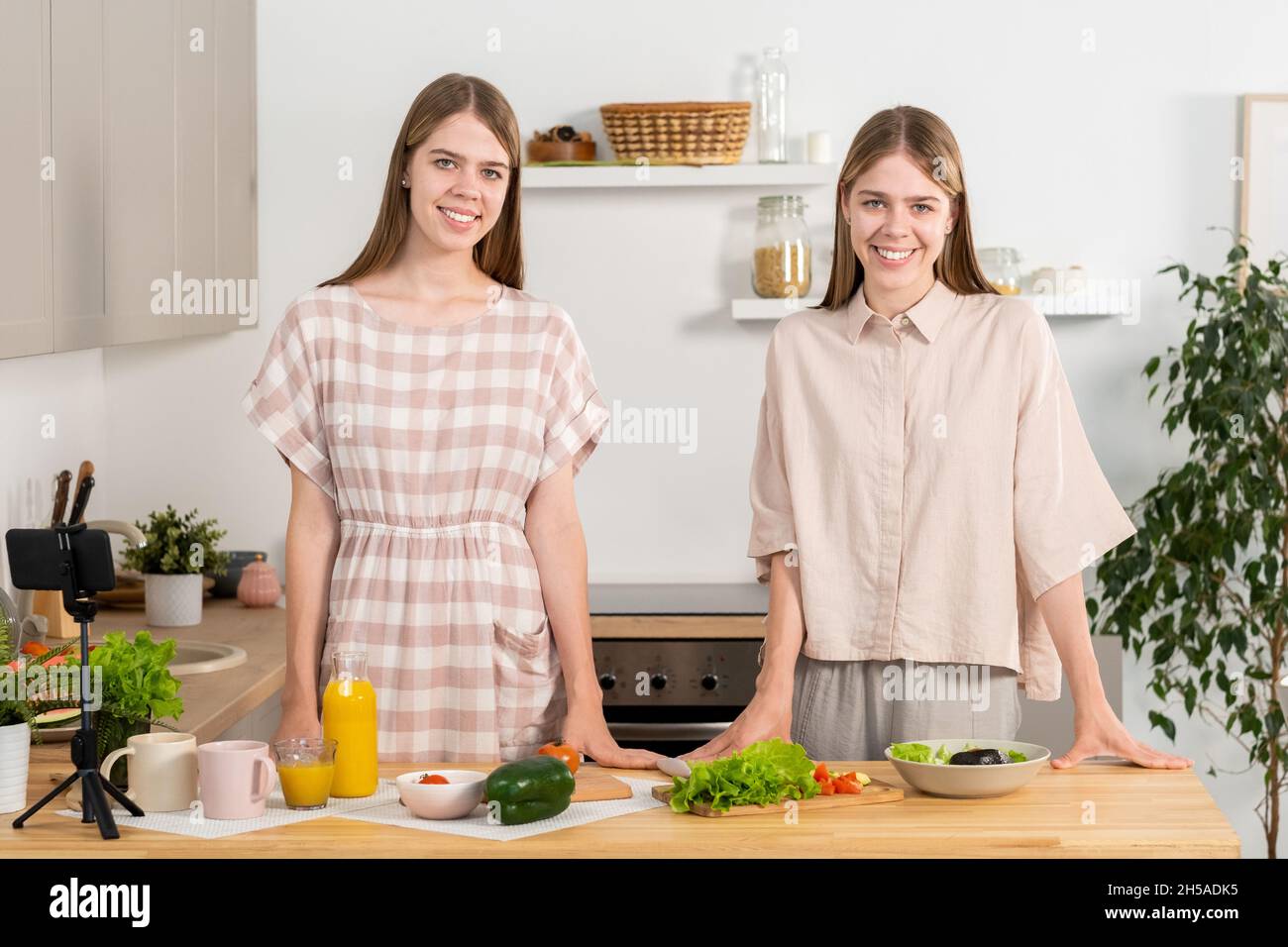 Glückliche junge Zwillingsschwestern in Casualwear, die am Küchentisch stehen und die Kamera betrachten, während sie vegetarisches Essen kochen Stockfoto