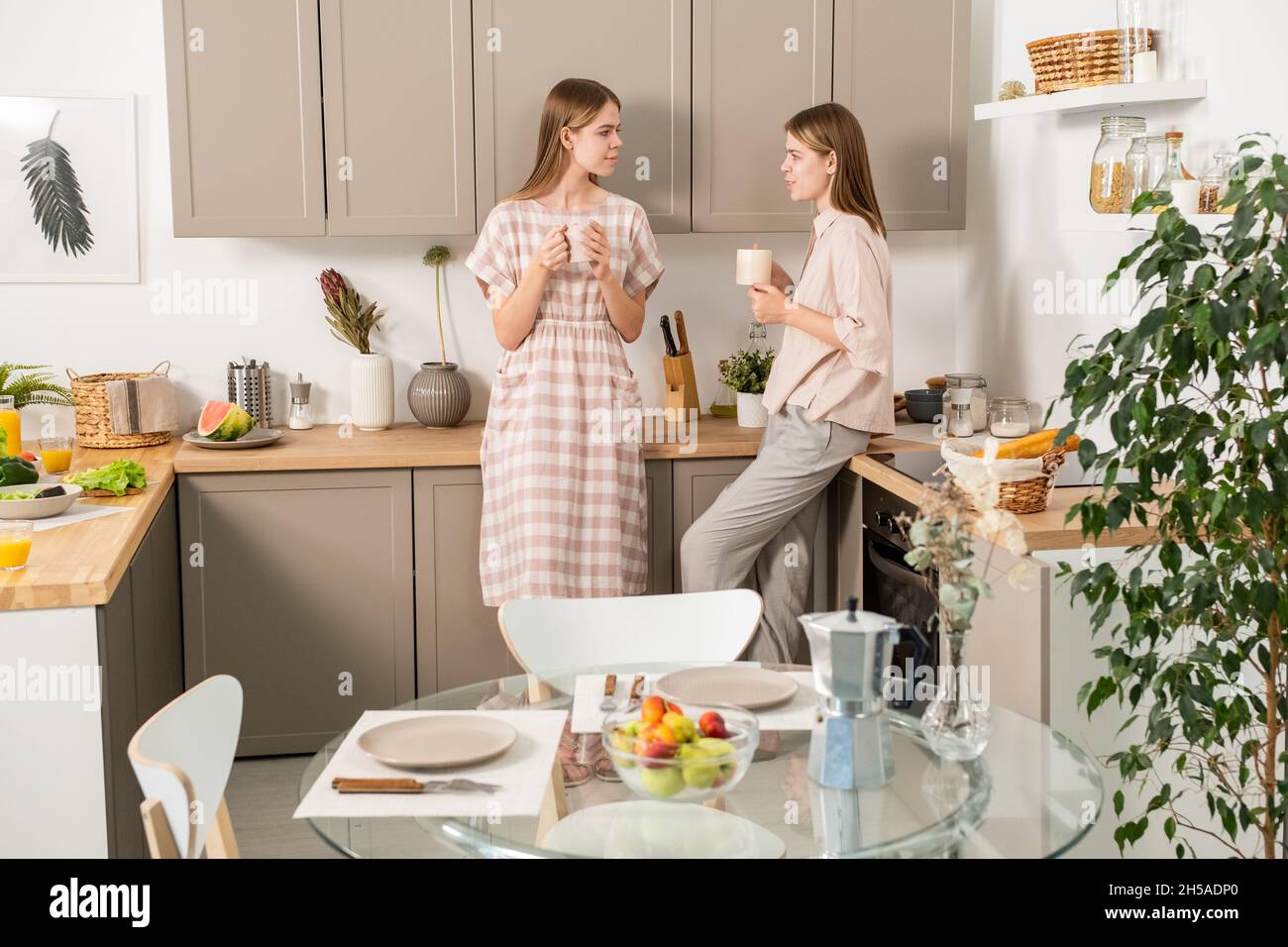 Zwei moderne Mädchen im Teenageralter in Casualwear diskutieren nach dem Frühstück über Pläne für ein Wochenende, während sie in der Küche einen Drink genießen Stockfoto
