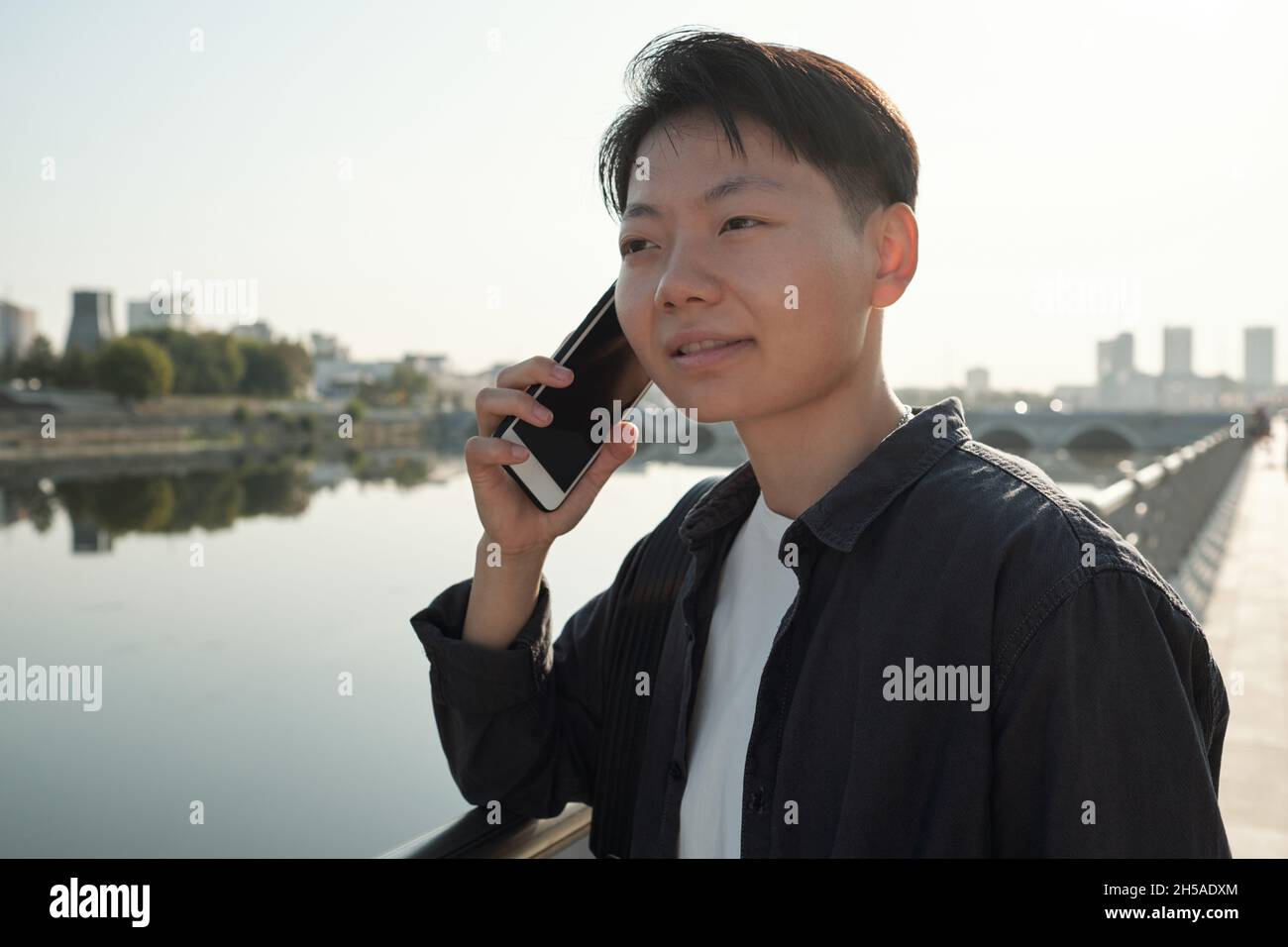 Hübsche chinesische Geschäftsagentin mit Smartphone am Ohr, die einen Kunden beim Stehen am Fluss konsultiert Stockfoto