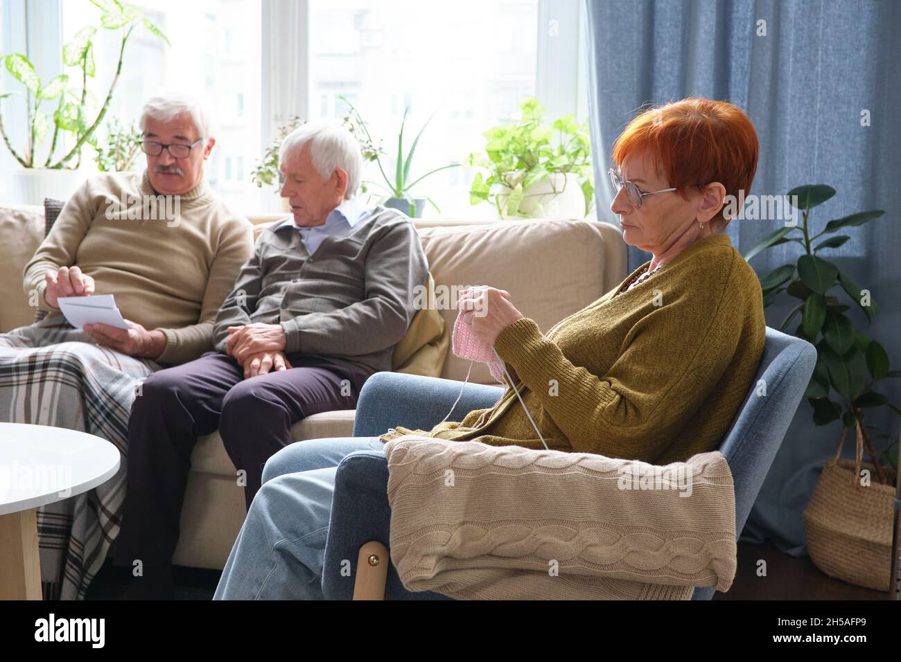 Eine ältere Frau sitzt im Sessel und strickt mit zwei älteren Männern, die zusammen auf dem Sofa im Hintergrund Fotos diskutieren Stockfoto