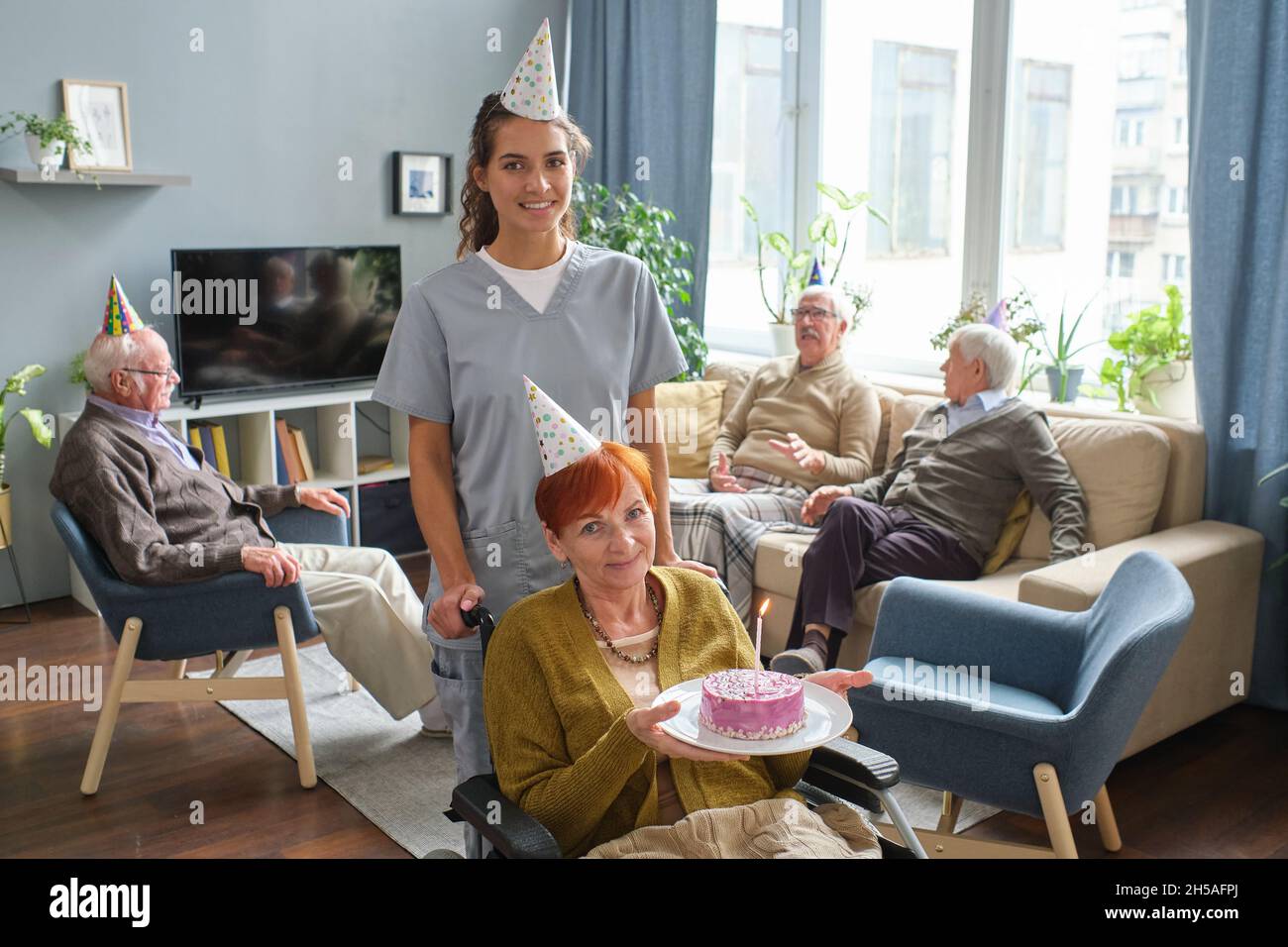 Porträt einer älteren Frau, die im Rollstuhl sitzt und die Kamera anlächelt und einen Geburtstagskuchen hält, während die Krankenschwester hinter ihr steht und im Pflegeheim Geburtstag feiert Stockfoto