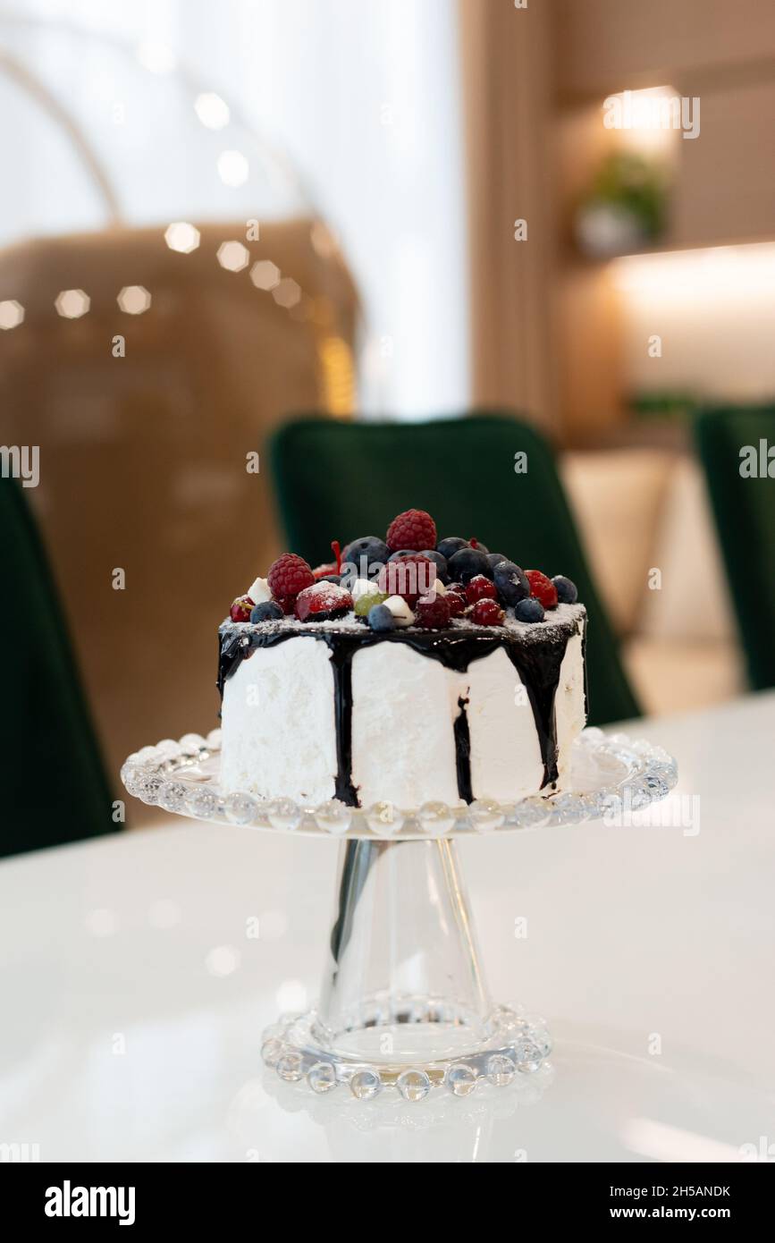 Gourmet-Kuchen mit Schokoladenüberzug und Beeren auf einem Glasständer auf einem weißen Tisch im modernen Interieur. Weicher, selektiver Fokus. Stockfoto