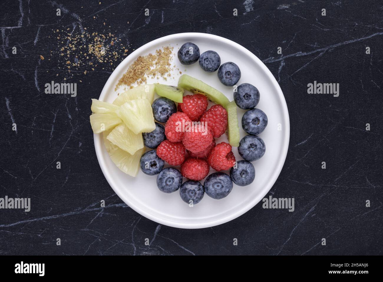 Weißer Teller mit frischen Beeren und Früchten auf schwarzem Marmor Stockfoto