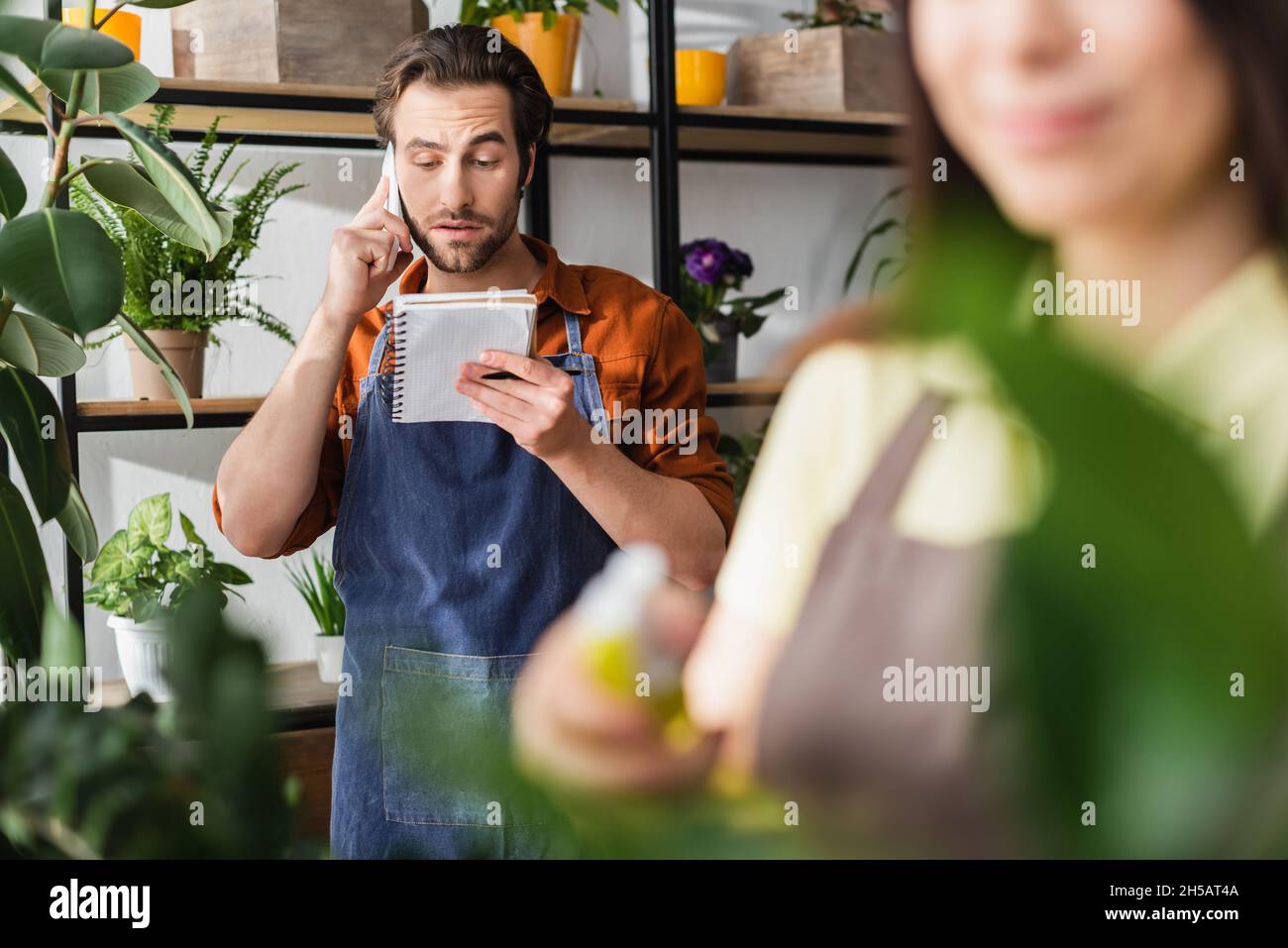 Junge Floristin, die am Handy sprach und Notebook in der Nähe von Pflanzen ansah, und verschwommener Kollege im Geschäft Stockfoto