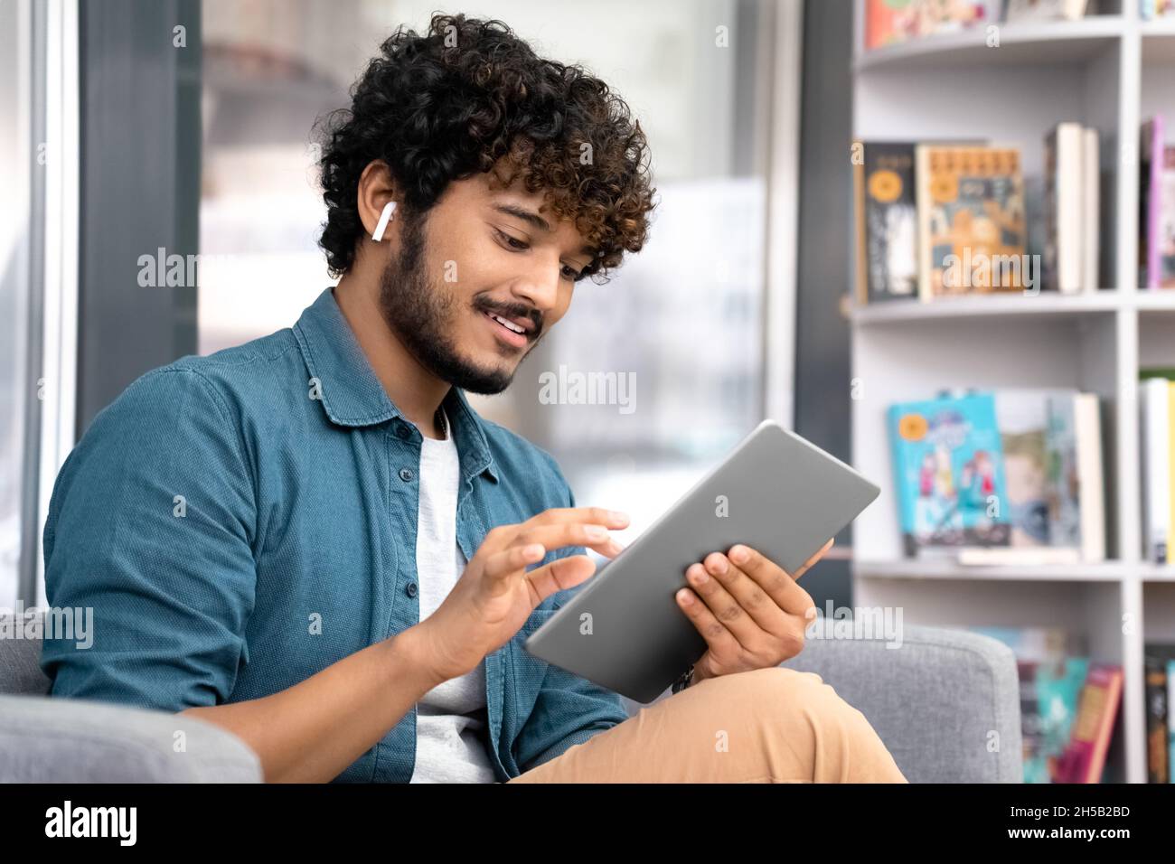 Junge Studentin in Ohrhörern mit einem mobilen Tablet, die auf einem Sessel in der Bibliothek sitzt Stockfoto