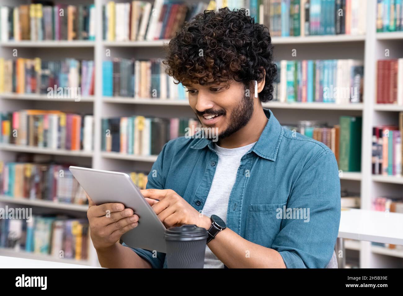 Junge indische Studenten mit Ohrhörern verwenden ein mobiles Tablet, das in der Bibliothek sitzt Stockfoto