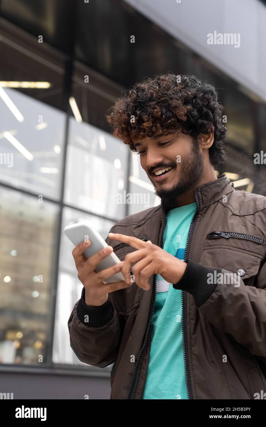 Glücklich lächelnder junger indischer Mann, der mit dem Mobiltelefon die Stadtstraße läuft Stockfoto