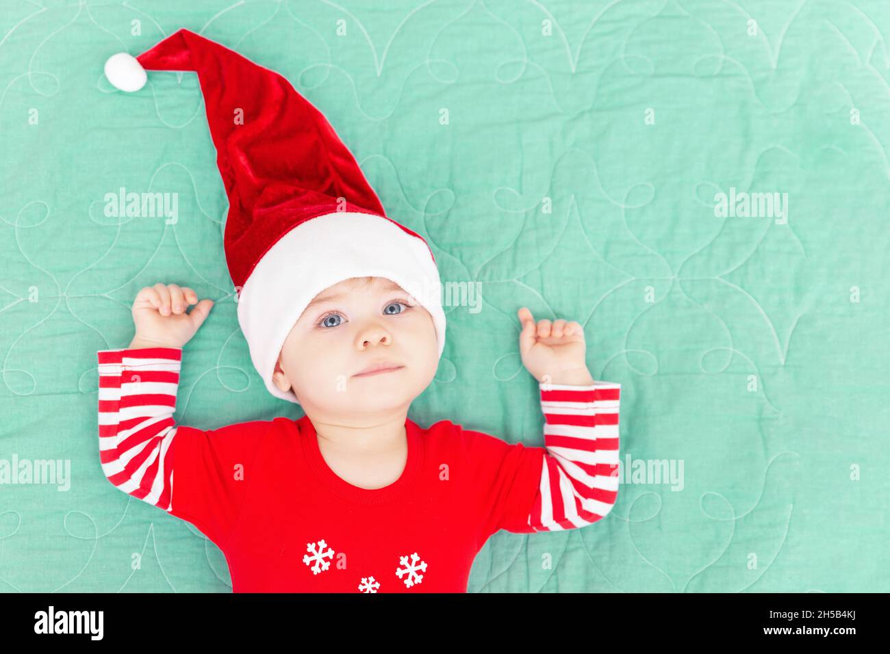 Lächelnder Kleinkind Junge liegt auf grünem Deckchen. Weihnachten Baby trägt großen roten Weihnachtsmann Hut Blick auf die Kamera. Neujahrskonzept. Draufsicht. Speicherplatz kopieren Stockfoto