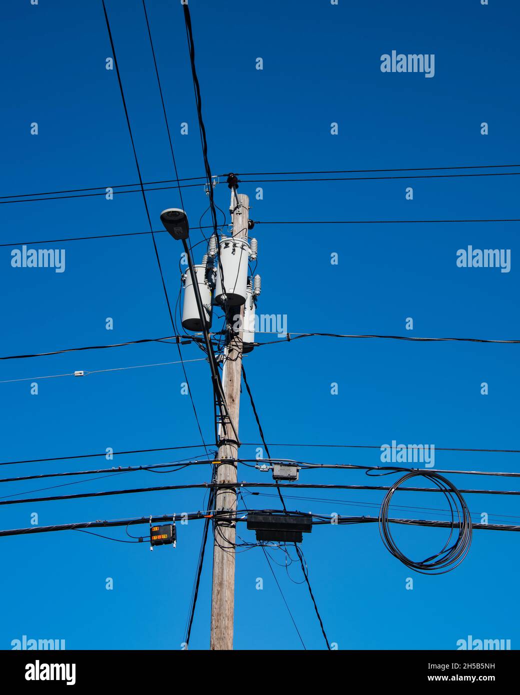 Ein Strommast mit Transformatoren, einer Straßenbeleuchtung, Verteilerkästen und Drähten und Kabeln mit einem tiefblauen Himmel Hintergrund. Stockfoto
