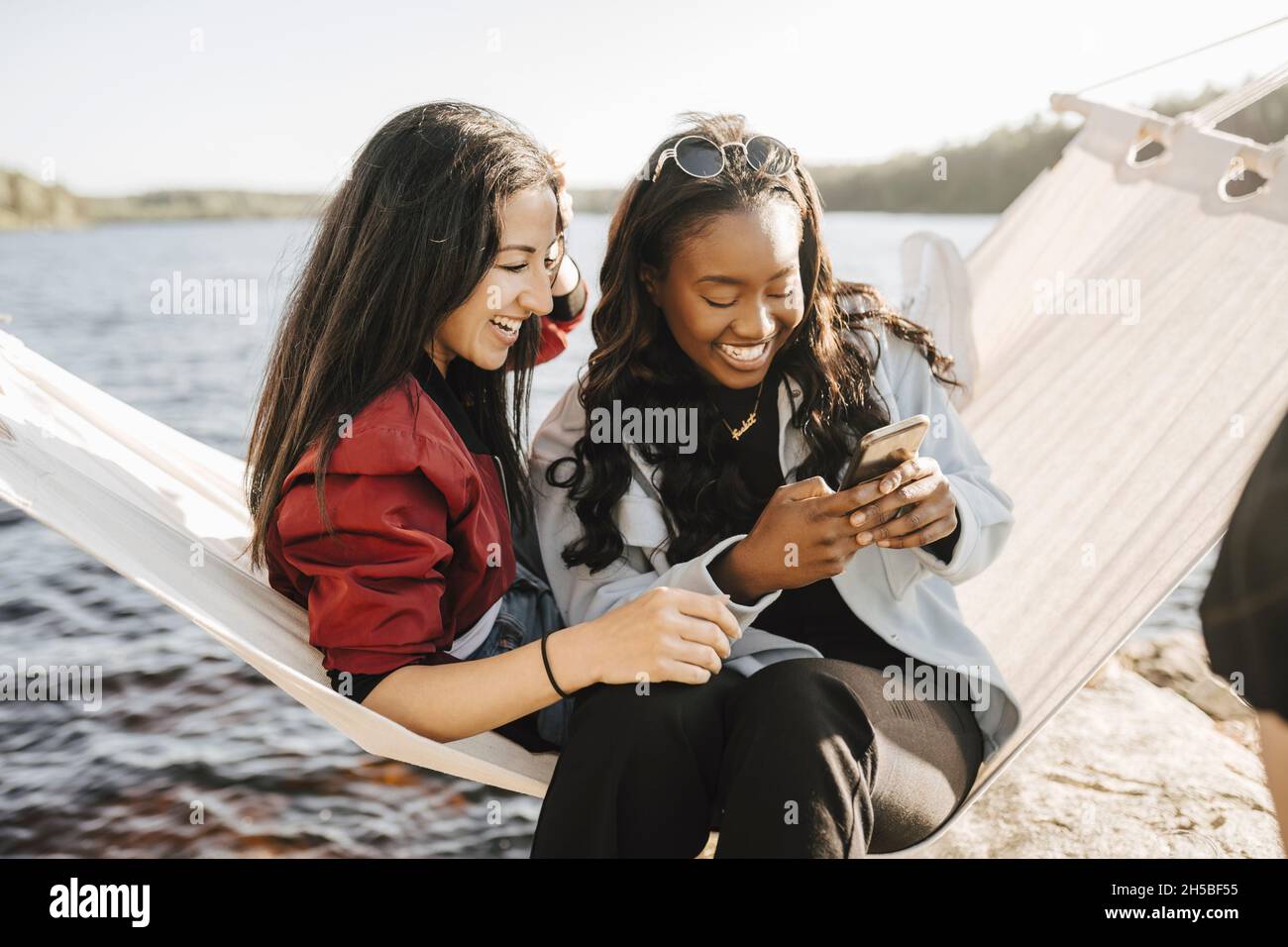 Glückliche weibliche Freunde, die Smartphone benutzen, während sie auf Hängematte sitzen Stockfoto